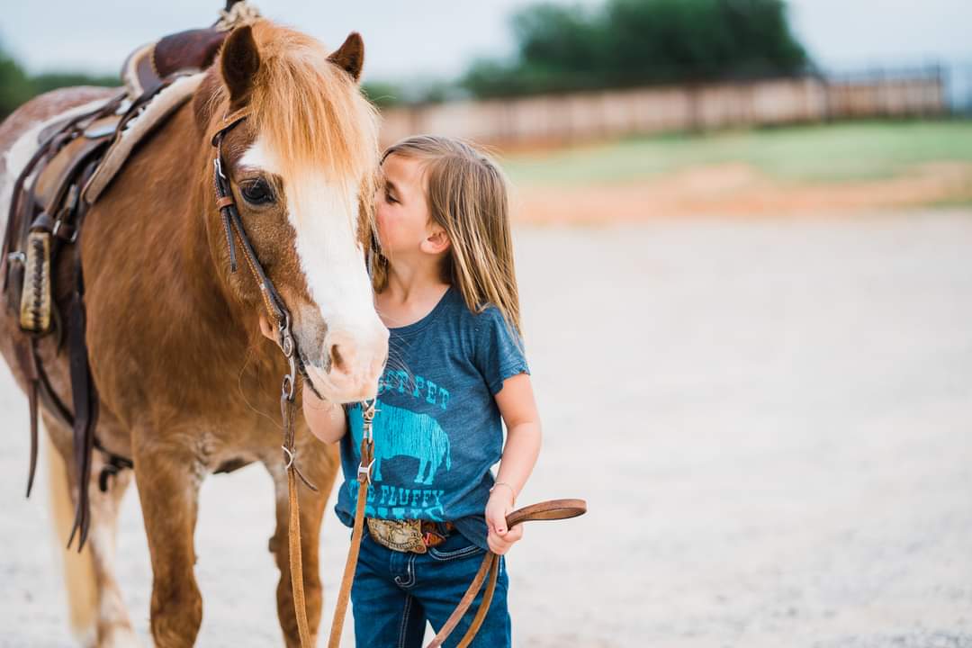 Young girl in a western graphic tee holding the reins and kissing her pony — a heartwarming back-to-school moment in true cowboy style
