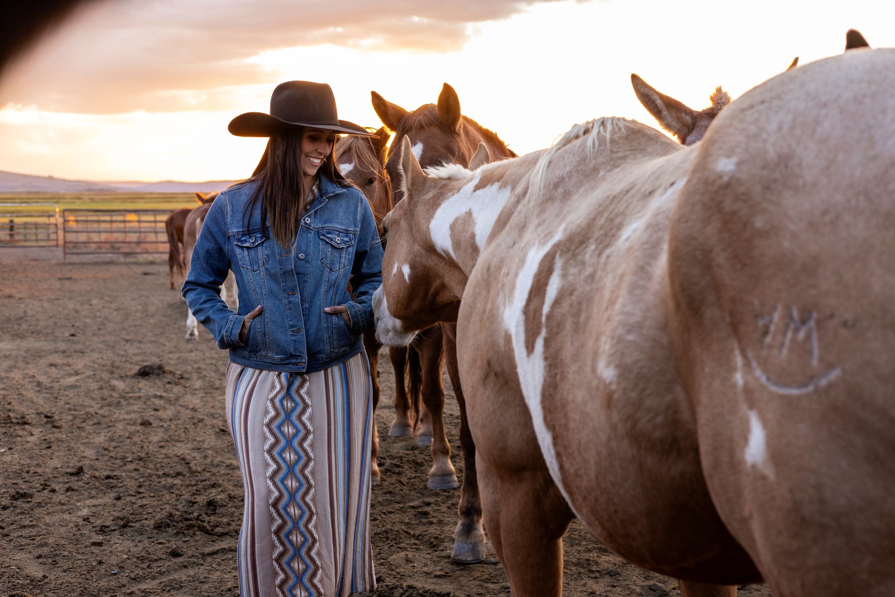 Woman in a denim jacket and patterned western maxi dress petting horses at sunset — a cozy fall western outfit perfect for cooler weather and ranch life
