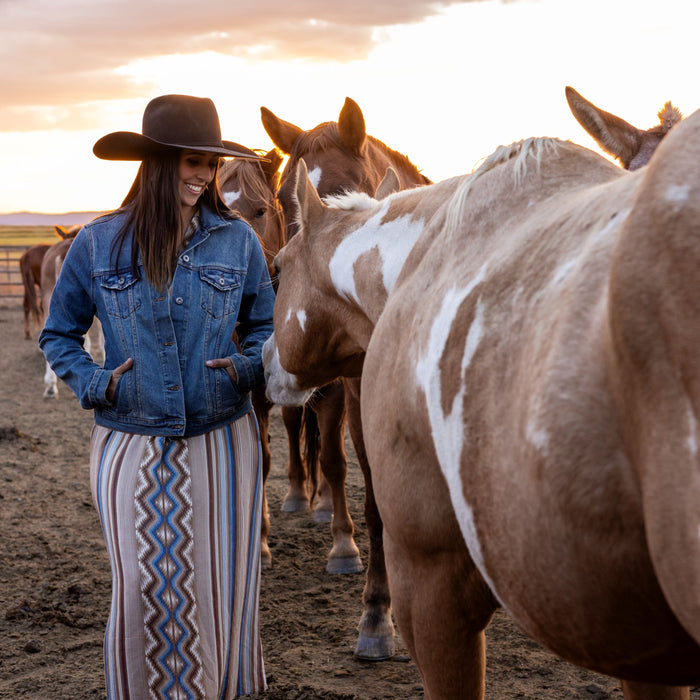Woman in a denim jacket and patterned western maxi dress petting horses at sunset — a cozy fall western outfit perfect for cooler weather and ranch life